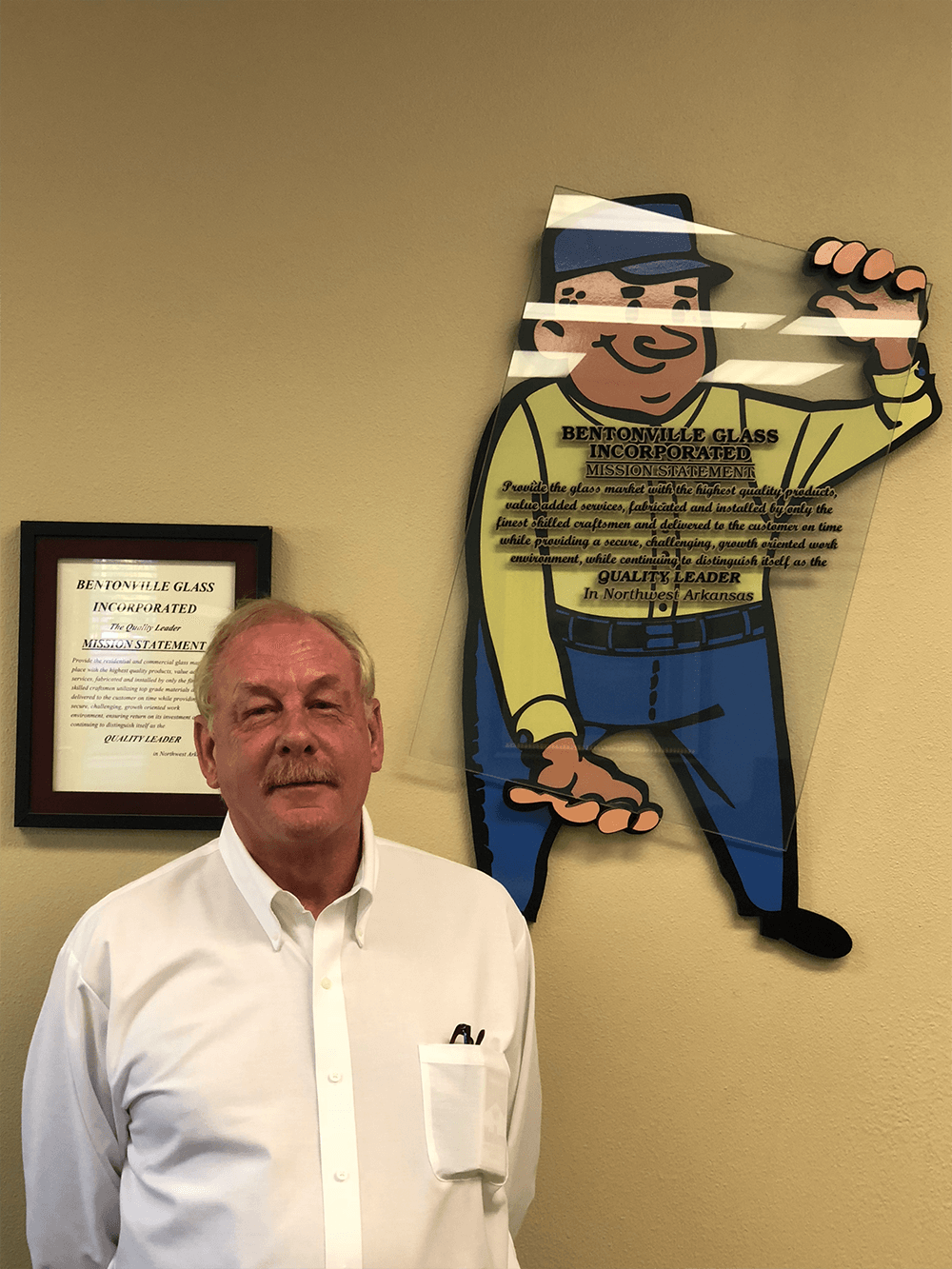 Man in white shirt standing beside a cartoon of a worker, a framed certificate hangs on the wall.