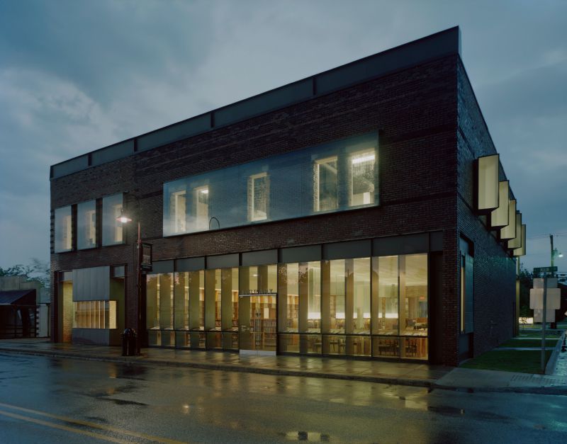 Brick building with large glass windows lit from inside, under a cloudy sky.