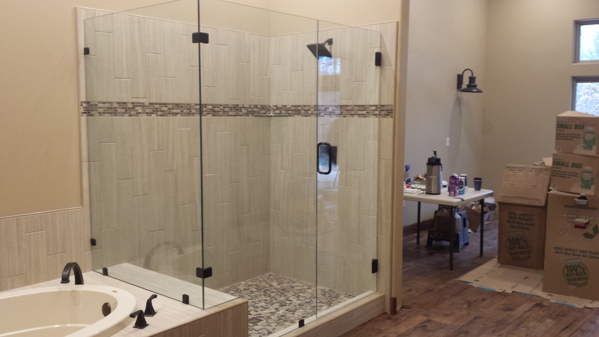 Glass shower enclosure with pebble floor, beige tiled walls, black fixtures, and hardwood flooring.