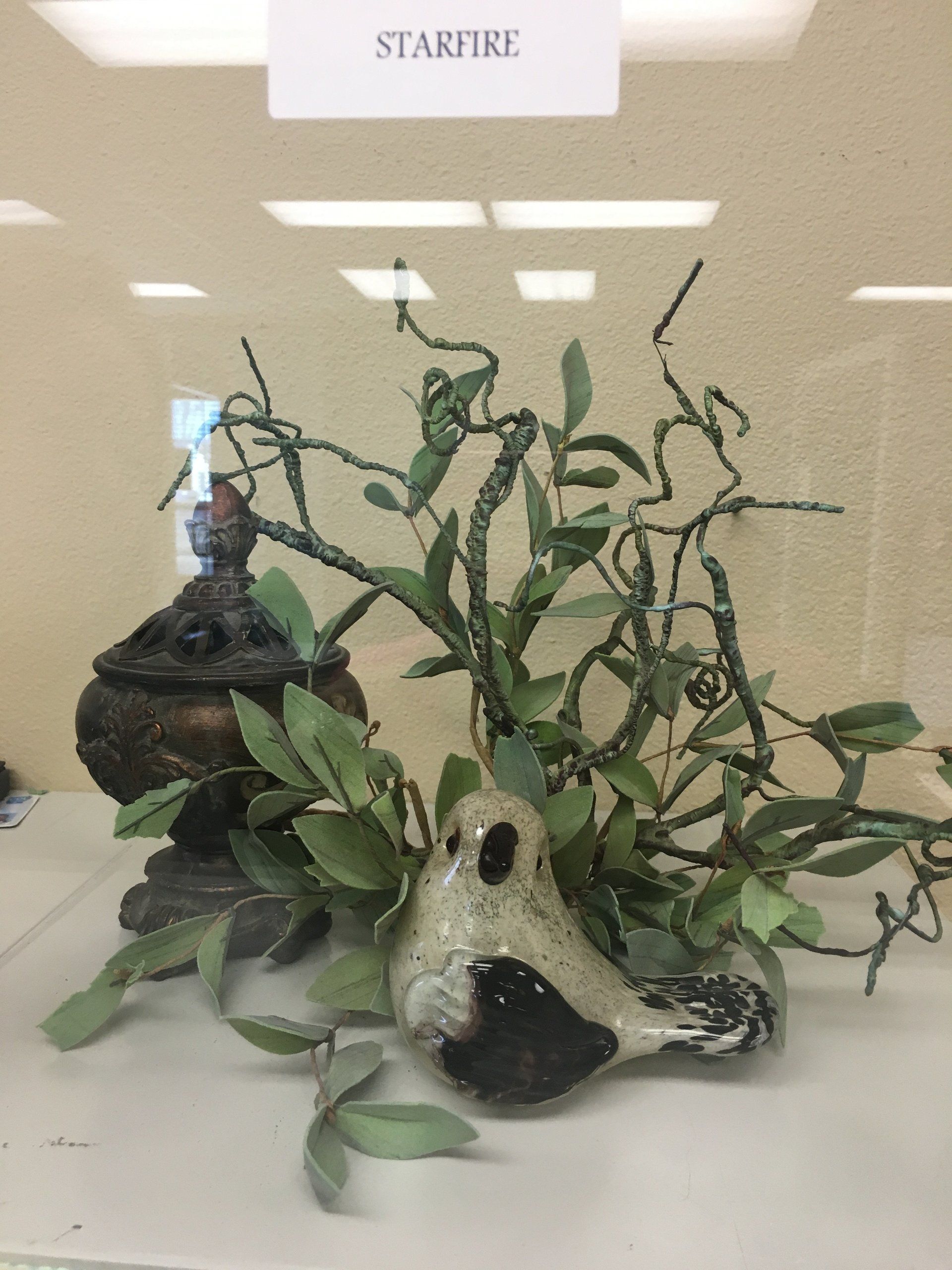 Display case with ceramic bird, decorative greenery, and a bronze-colored container, labeled 