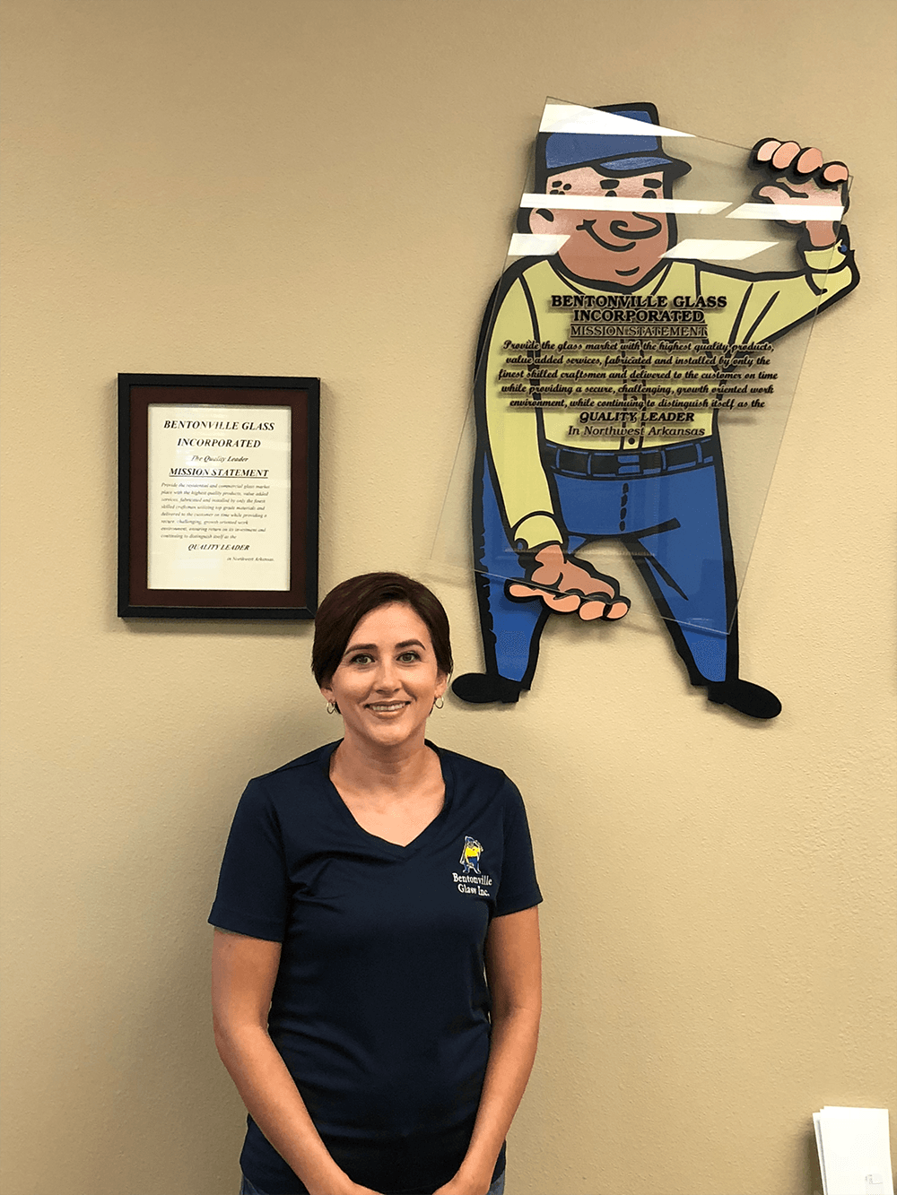 Woman in blue shirt smiling in front of a wall with a cartoon character and framed document.