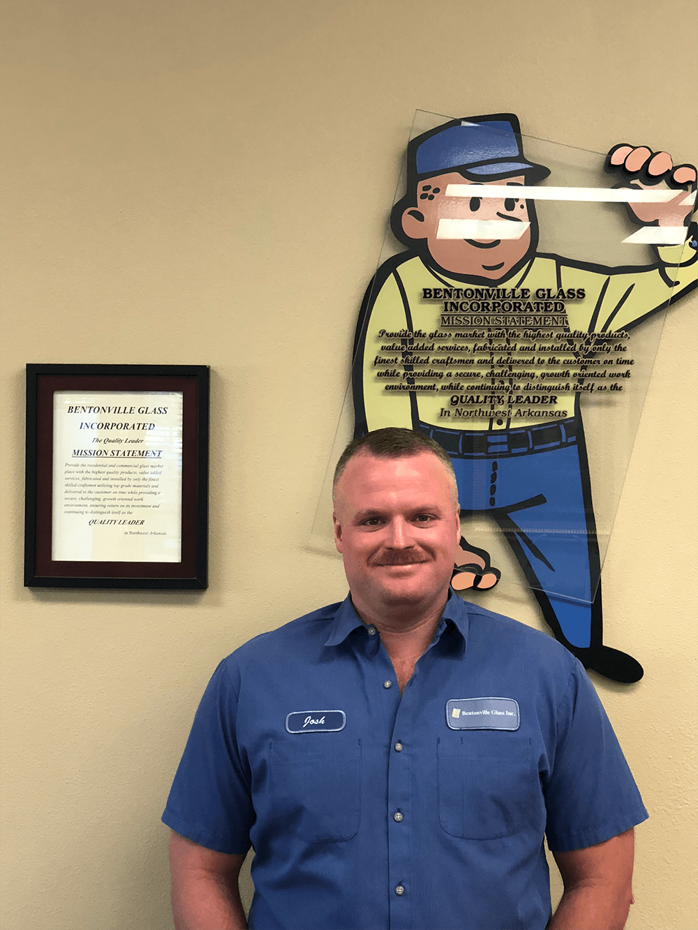 Man in blue uniform stands in front of a cartoon figure; a framed document hangs on the wall.