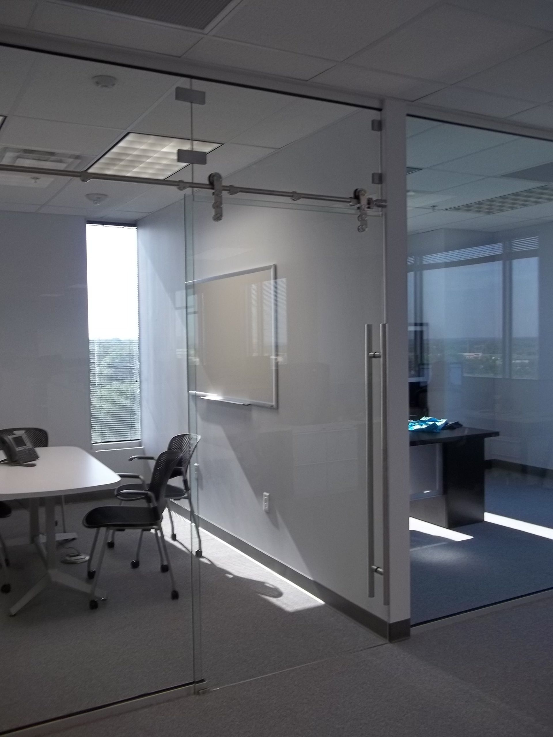 Glass-walled office space with a conference table and a desk. Sunlight streams through a window.