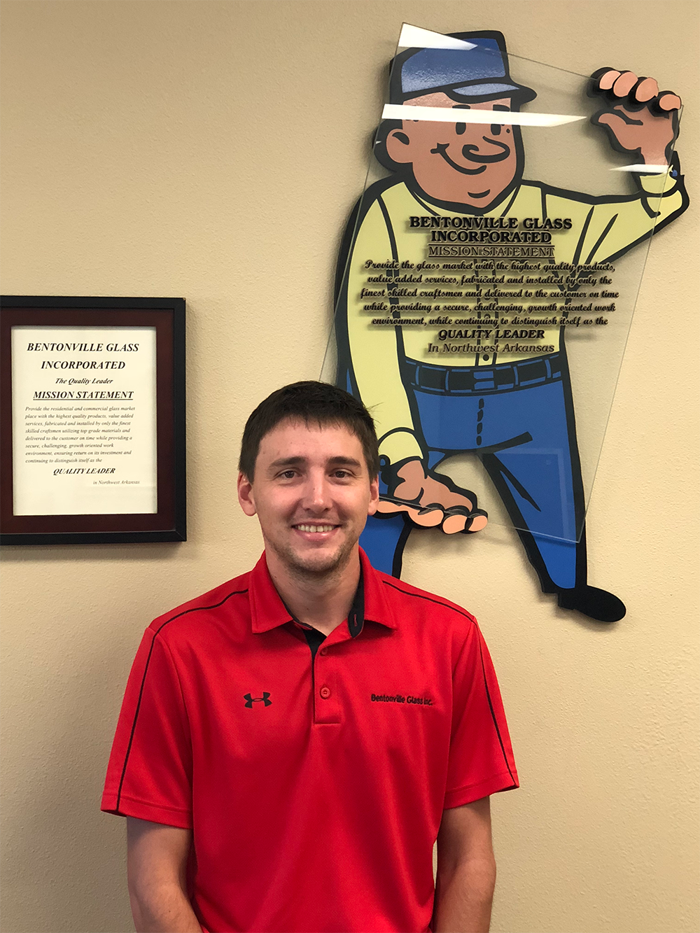 Man in red polo shirt smiles in front of a cartoon mascot and framed certificate.