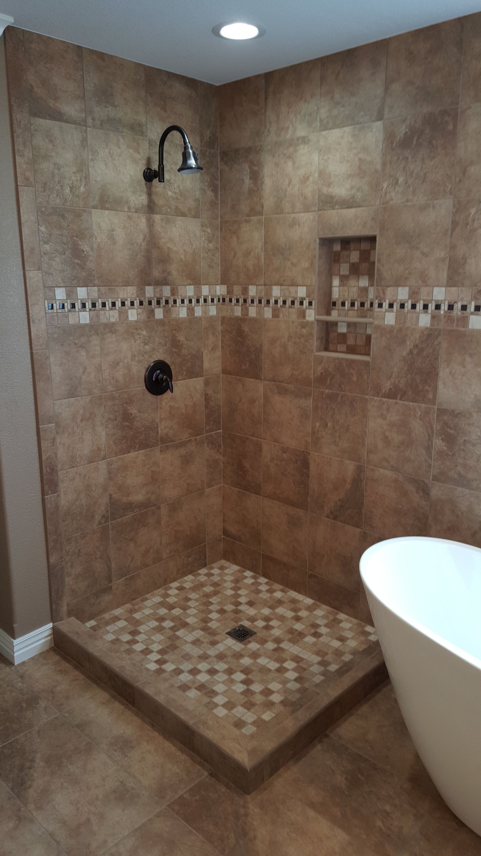 Corner shower with brown tile, mosaic floor, dark fixtures. White tub on right.