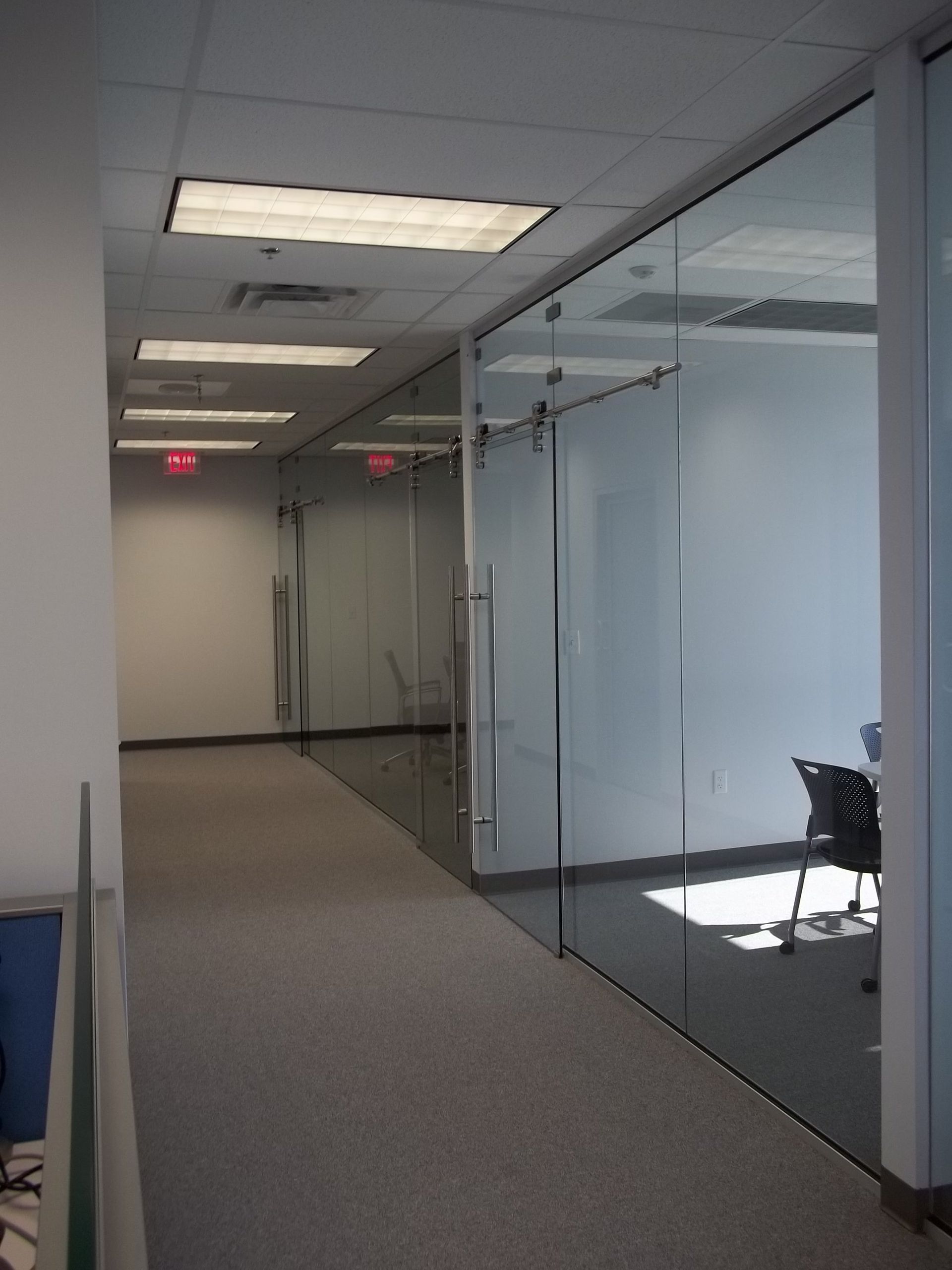 Office hallway with glass-walled rooms, carpeted floor, and fluorescent lighting.