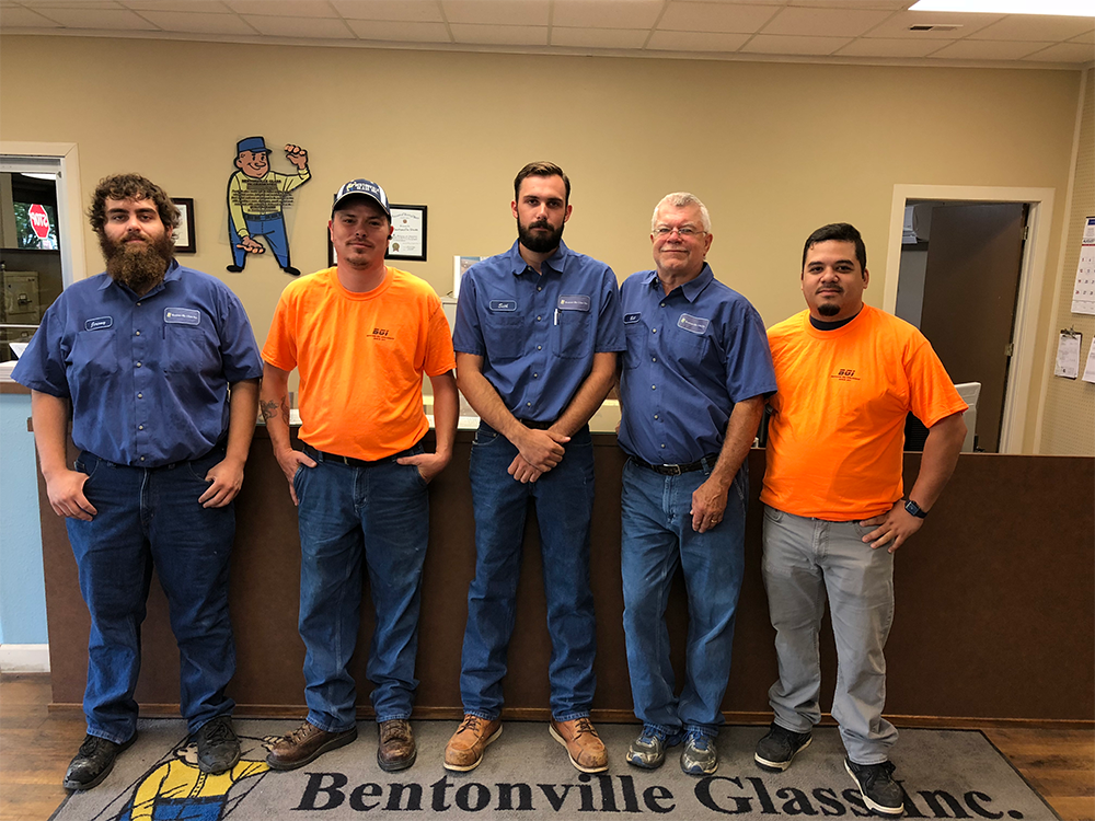 Five employees of Bentonville Glass Inc. standing inside. Two wear orange shirts, three in blue.