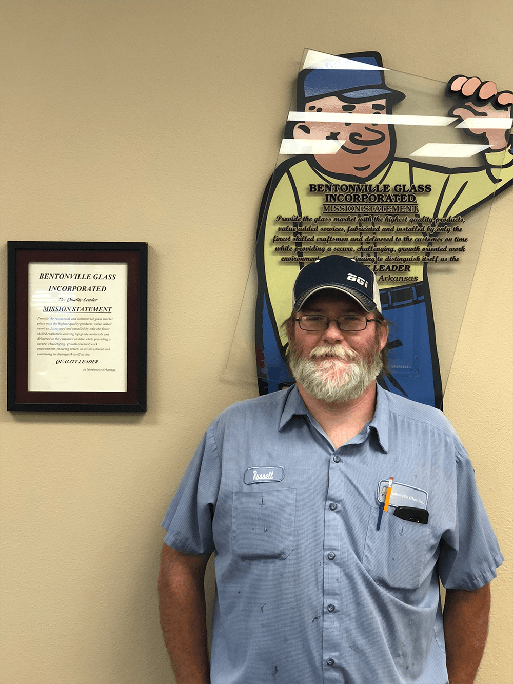 Man with beard, blue shirt, and hat stands next to a wall with a framed document and a cartoon figure.