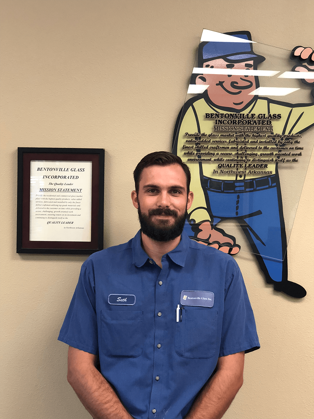 Man with a beard wearing blue shirt, standing in front of a wall with a framed document and a cartoon character.
