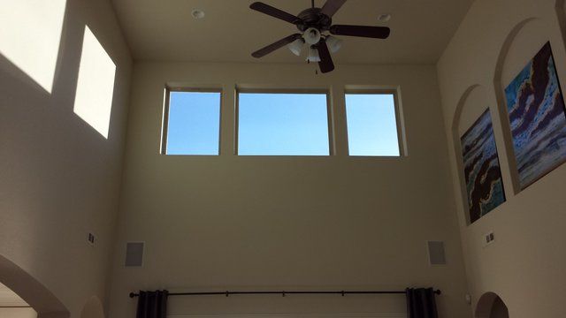 High-ceilinged living room with three windows against a blue sky. Artwork on walls, ceiling fan.