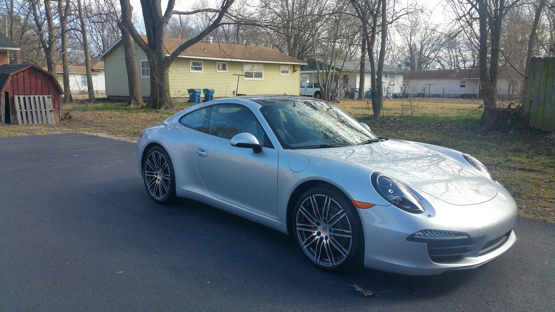 Silver Porsche parked on a paved driveway in a suburban neighborhood, with trees and houses in the background.