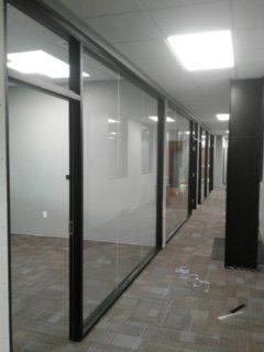 Office hallway with glass-walled rooms, black frames, beige carpet, and overhead lighting.