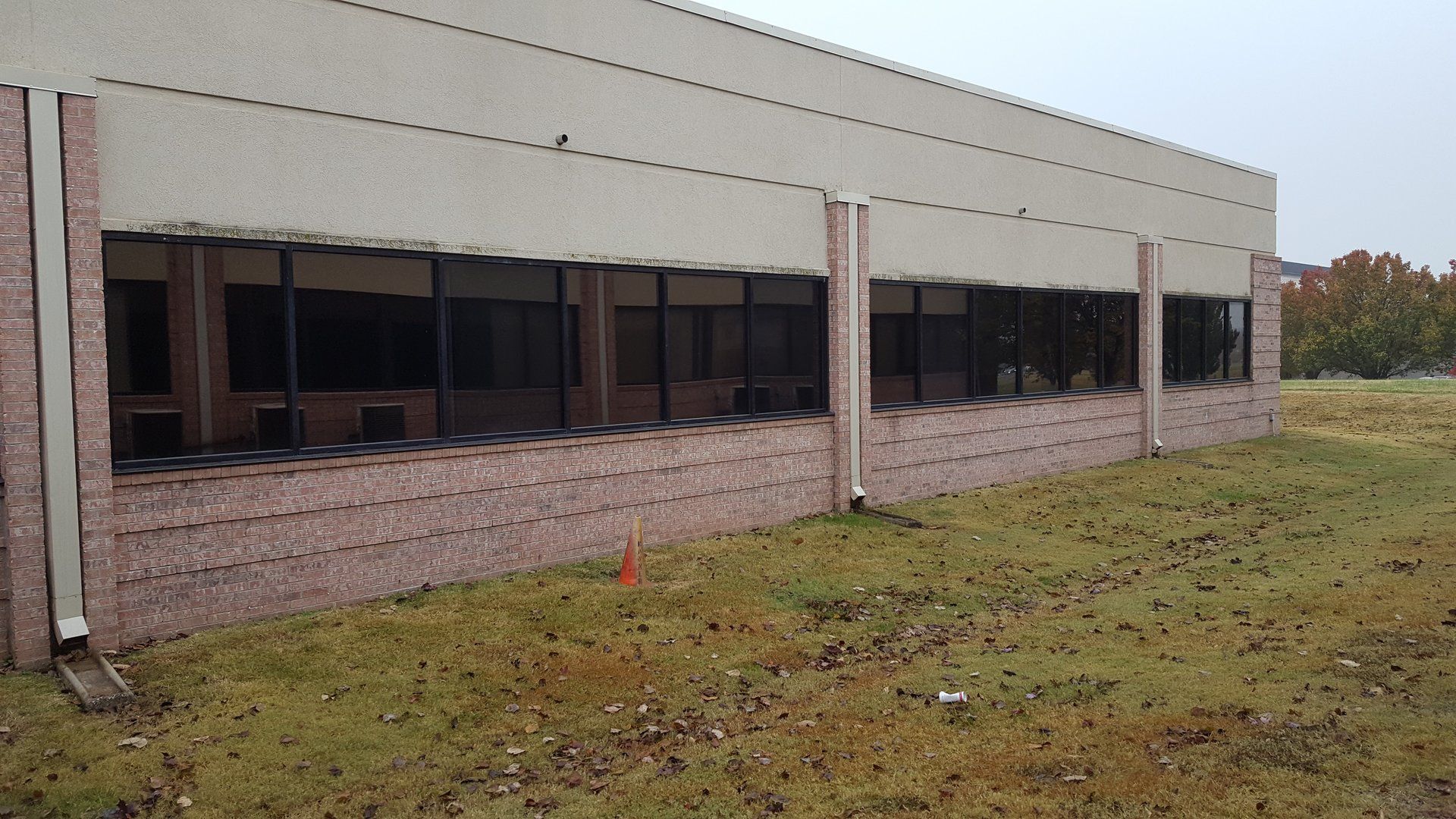 Long brick building with dark windows, brown and tan exterior, set in a grassy field.