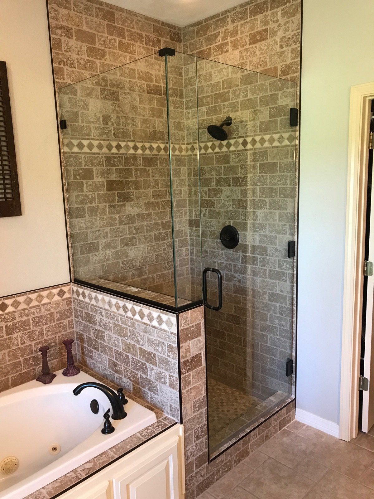 Corner shower with glass door and bronze fixtures, beside a jacuzzi tub with beige brick tile.