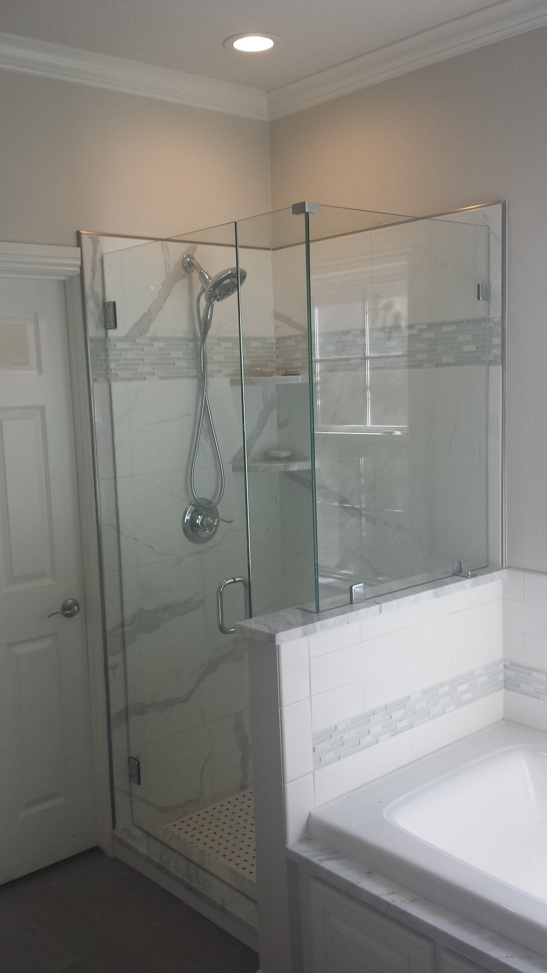 Glass shower in a modern bathroom, next to a white bathtub. White tile and gray walls.