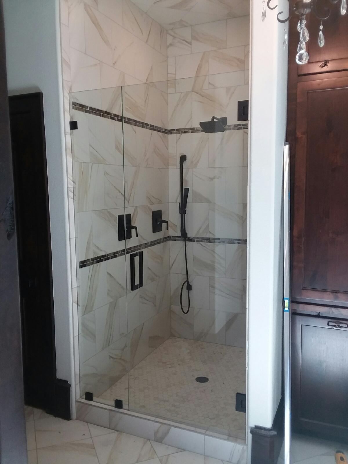 A modern, glass-walled shower in a tiled bathroom. Black fixtures, white marble tile, and a dark brown cabinet.