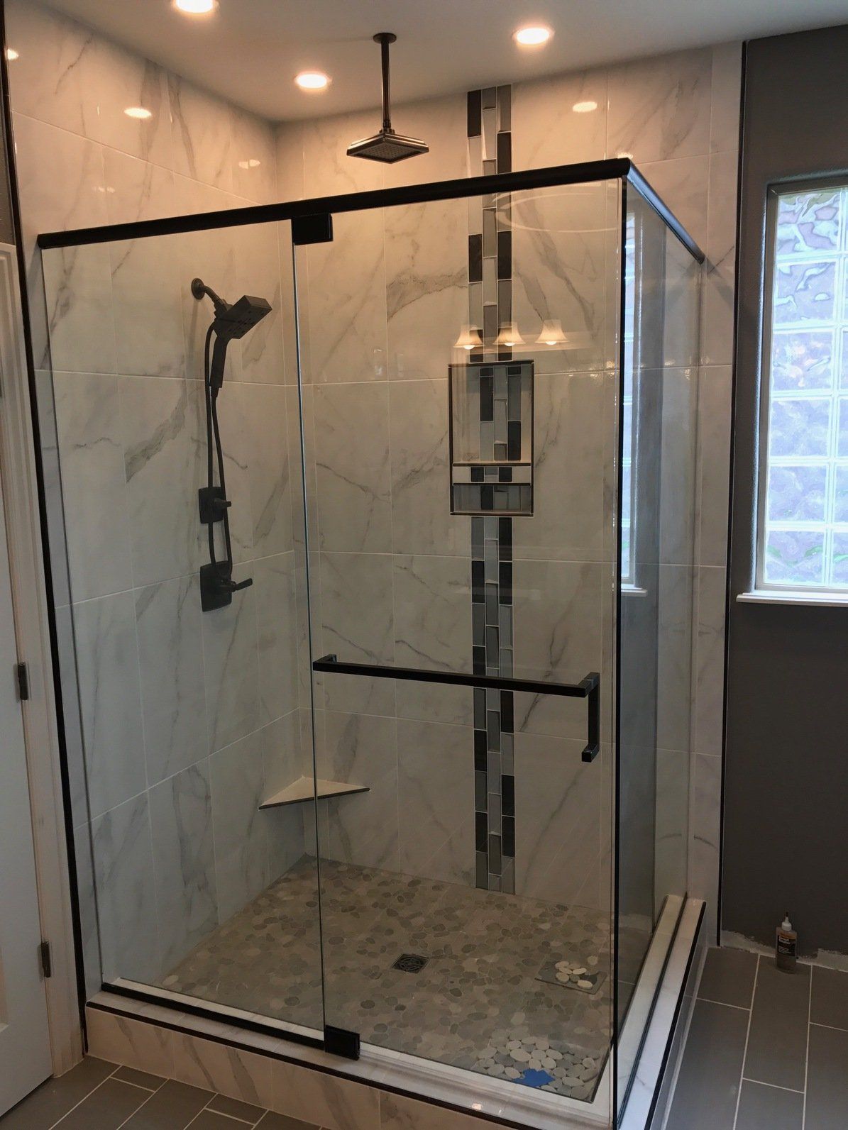 Modern bathroom with a glass-enclosed shower, marble-look tiles, dark fixtures, and a pebble floor.