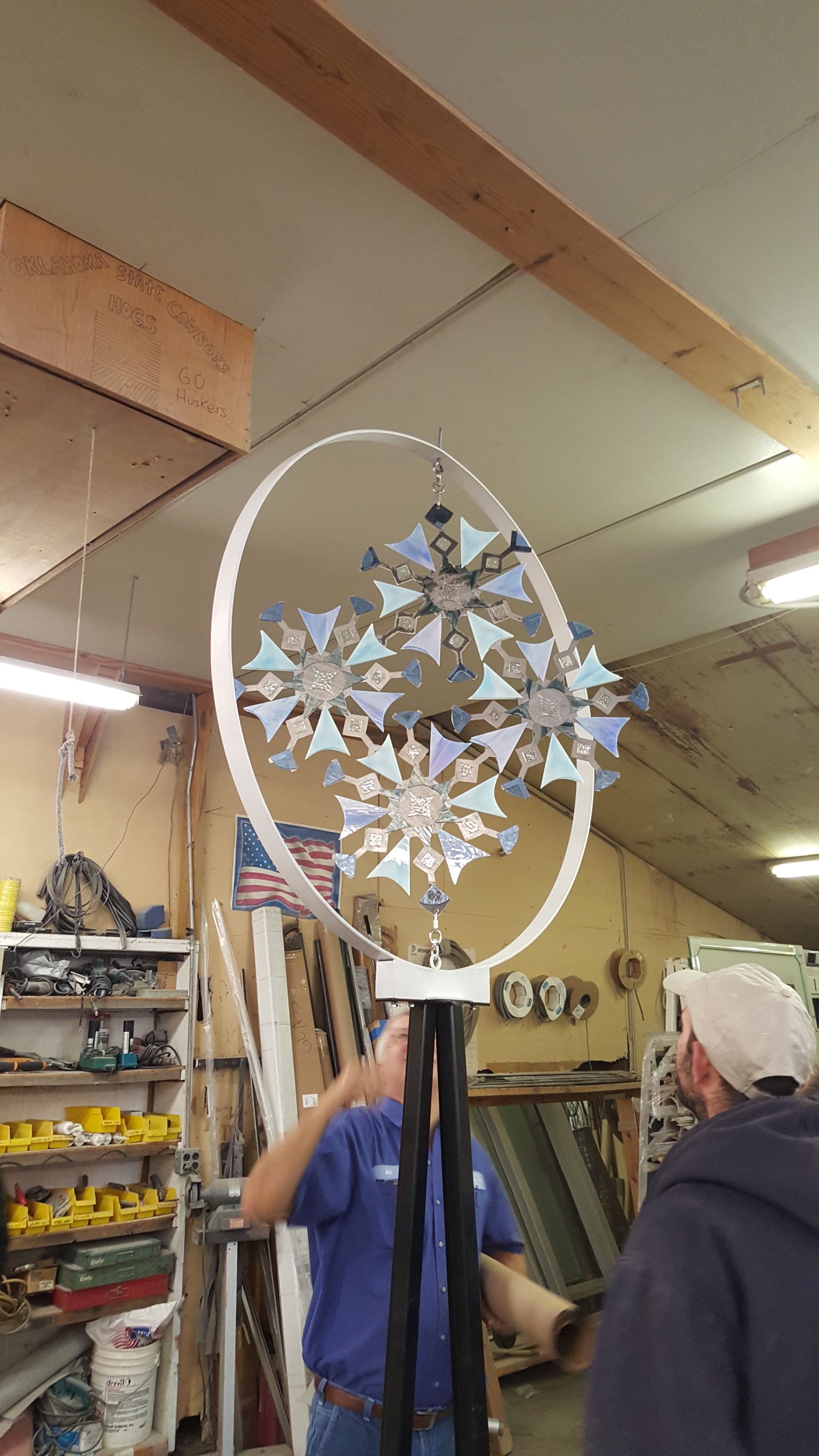 Two men assemble a metal wind spinner with a white ring and flower-like elements inside a workshop.
