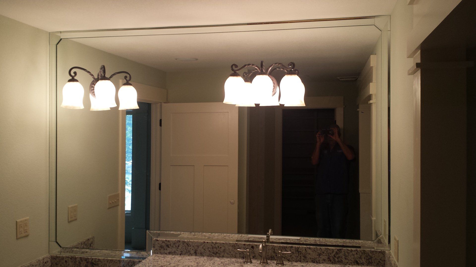 Bathroom mirror reflects lights, a door, and a person taking a photo. Granite countertop and light fixtures.