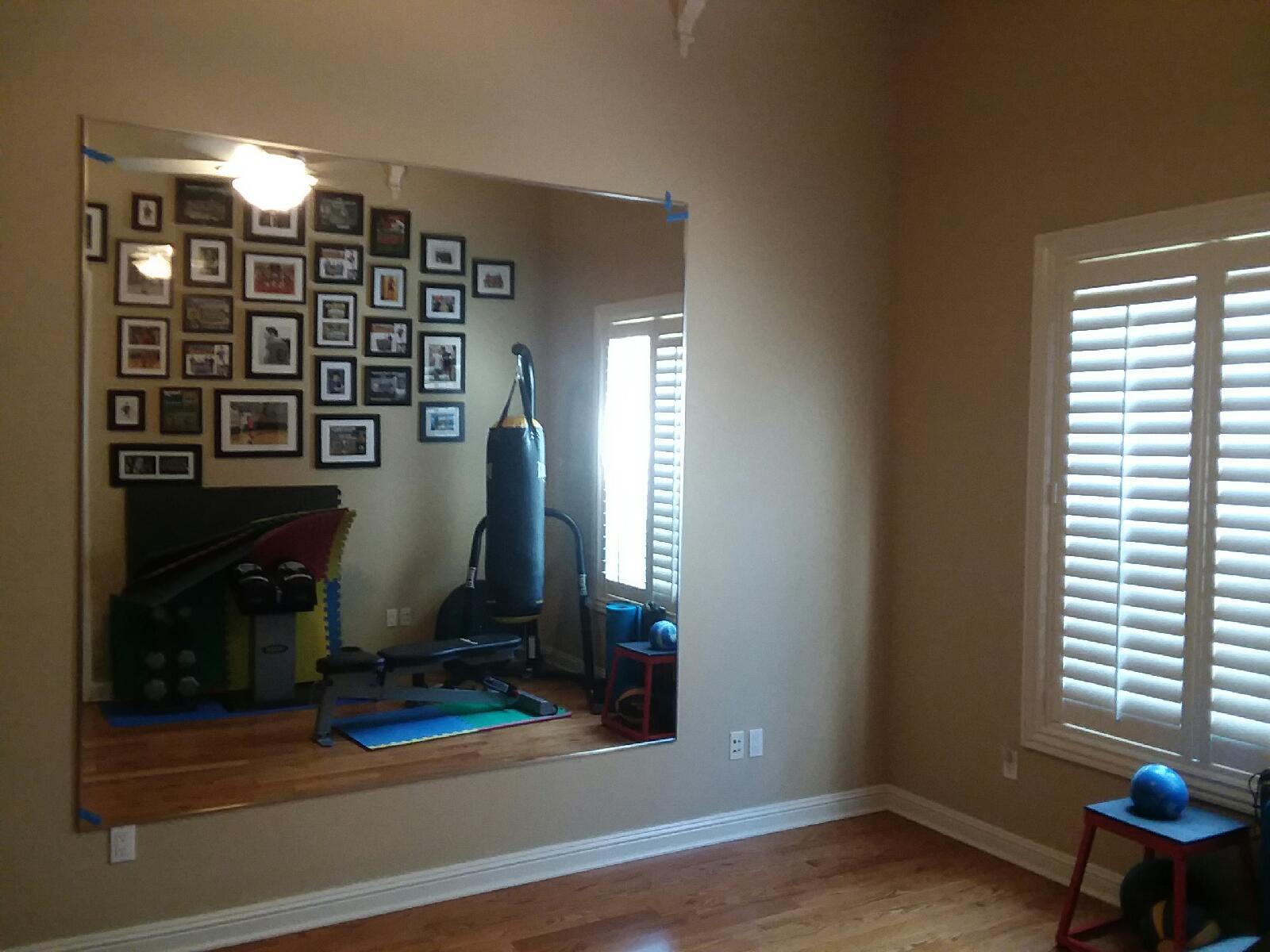 Large wall mirror reflecting a home gym with exercise equipment and a gallery wall. Window with shutters.
