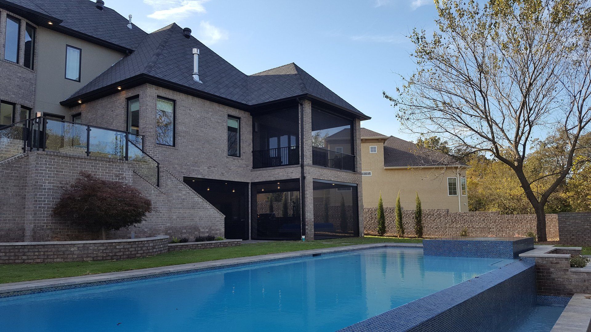 Back view of a large house with a pool. Beige brick, black screened porch, blue water. Sunny day.