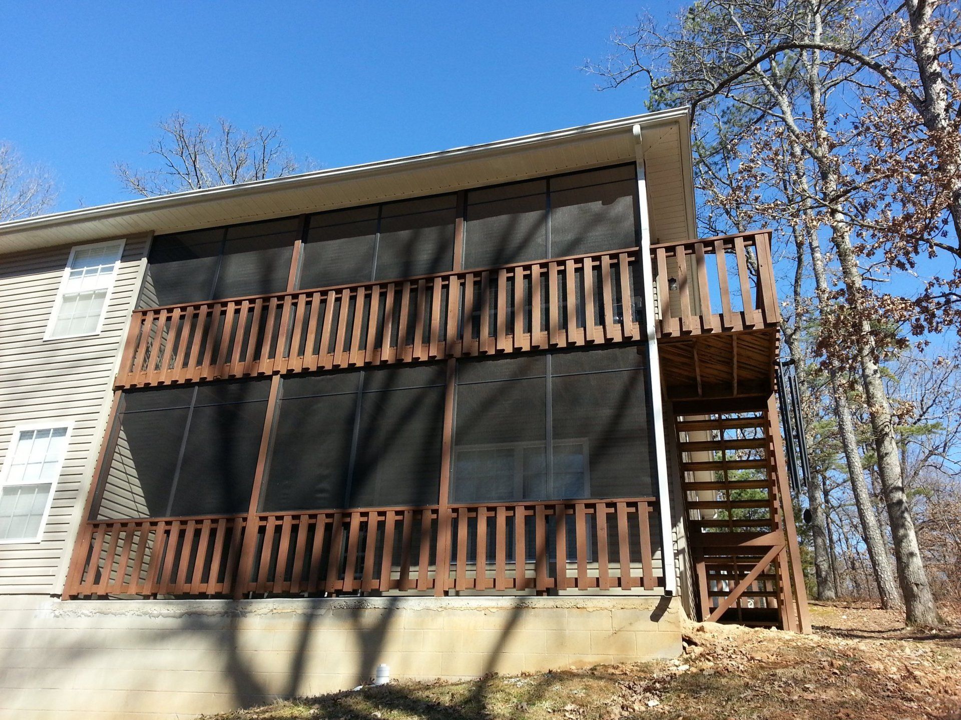 Two-story screened porch with brown railing, side stairs, and beige house against blue sky in wooded setting.