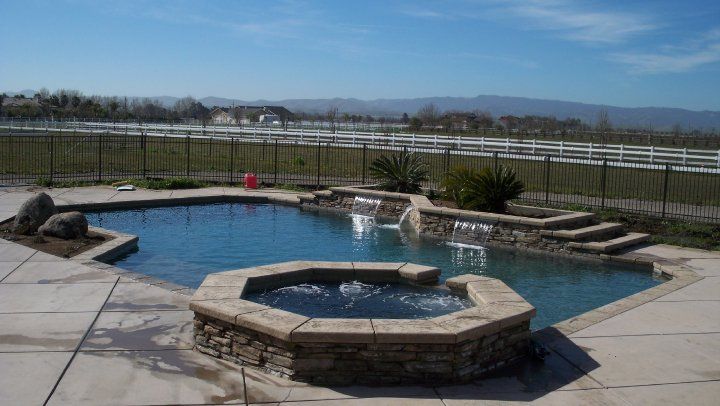 Pool and spa with waterfalls, surrounded by a paved patio. Sunny day with mountains in the background.