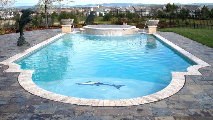 Swimming pool with light blue water, surrounded by stone patio. Spa with waterfall in the background, statue of dolphin.