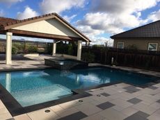 Pool and spa in a backyard patio with a covered seating area under a cloudy sky.