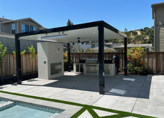 Outdoor kitchen with a black pergola, grill, fireplace, and a swimming pool in the background.