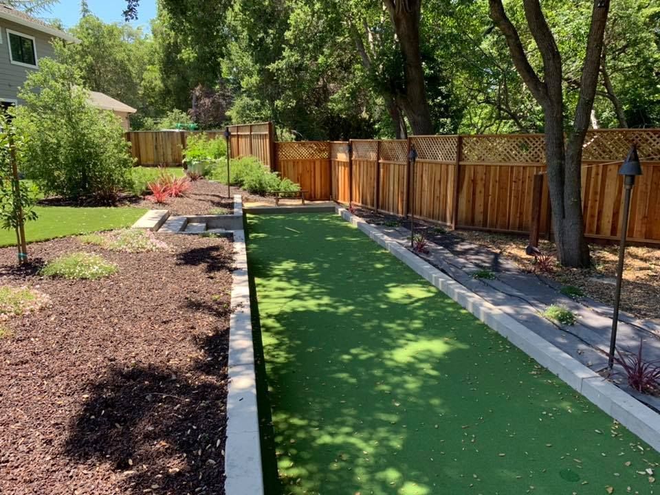 A backyard bocce ball court with green turf, bordered by concrete and wood-fenced.  Trees and shrubs surround the court.