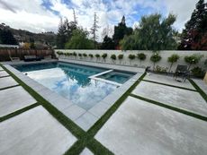 Swimming pool with surrounding concrete and turf, bordered by small trees and a wall, overcast sky.