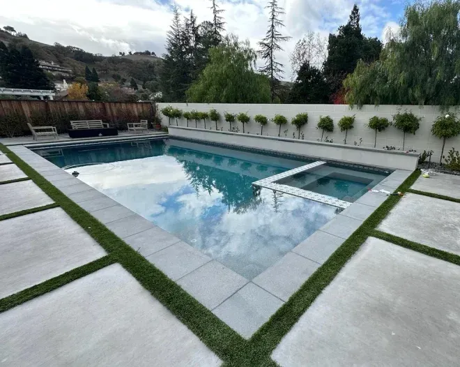 Rectangular pool and spa with blue water, surrounded by concrete and grass squares, against a hillside.