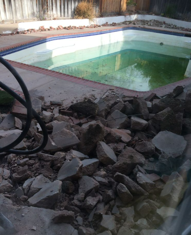 A dirty, algae-filled swimming pool with broken concrete debris in the foreground.