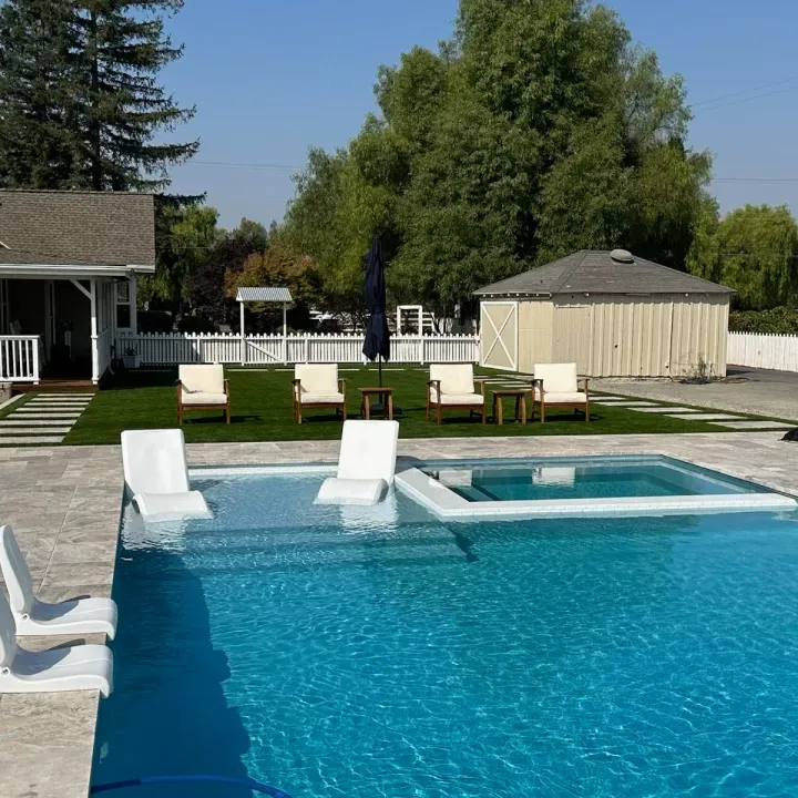 Poolside scene with pool, spa, lounge chairs, green lawn, beige building, blue umbrella, sunny day.