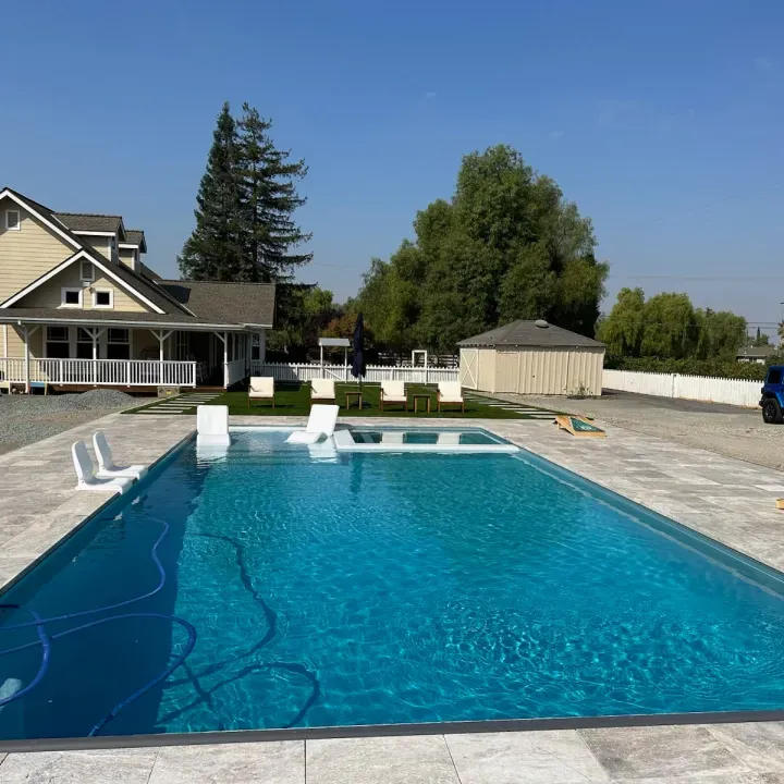 A rectangular swimming pool with blue water next to a light-colored house under a clear, blue sky.