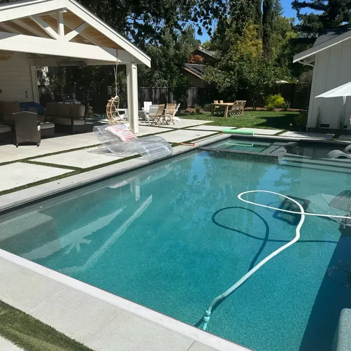 Pool area with turquoise water, a covered patio, and outdoor furniture on a sunny day.