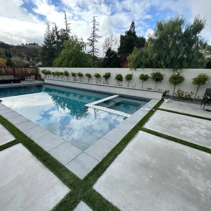 Pool area with rectangular pool, hot tub, concrete patio, and trees under a cloudy sky.