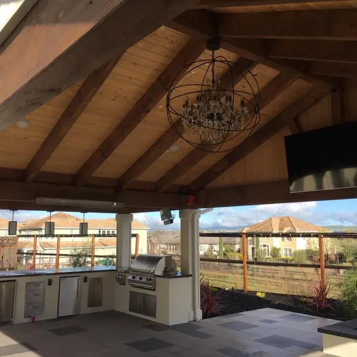 Outdoor kitchen with a grill, cabinets, and a chandelier under a wooden roof.