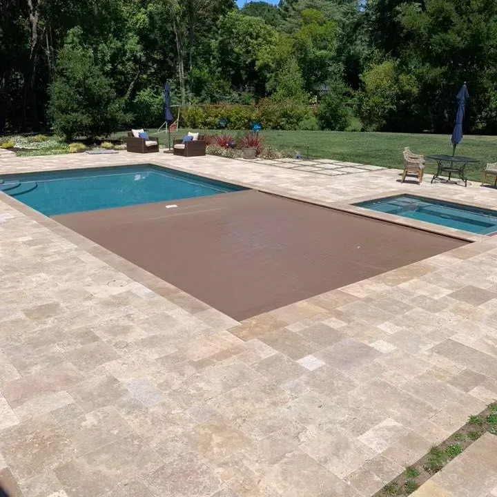 Pool with a brown cover, spa, and stone patio surrounded by grass and trees.