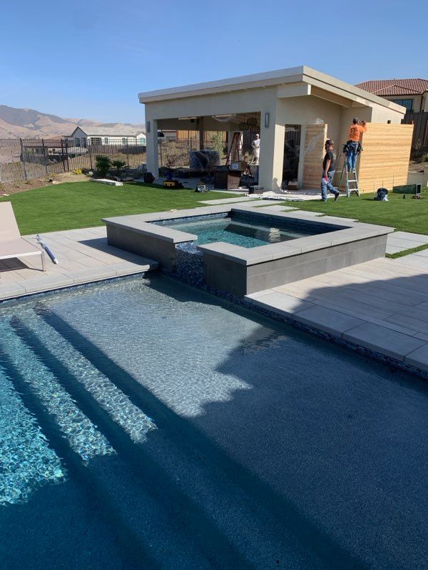 Pool with spa, pavilion, and lawn in a sunny backyard.  Man works near the pavilion. Blue water.