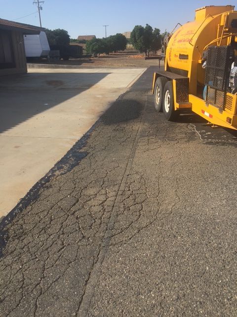 Yellow asphalt sealing trailer on a cracked asphalt surface. Sunlight, trees, and buildings in background.