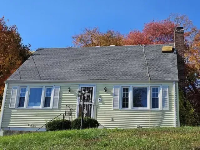A house with a gray roof and white shutters is sitting on top of a grassy hill.