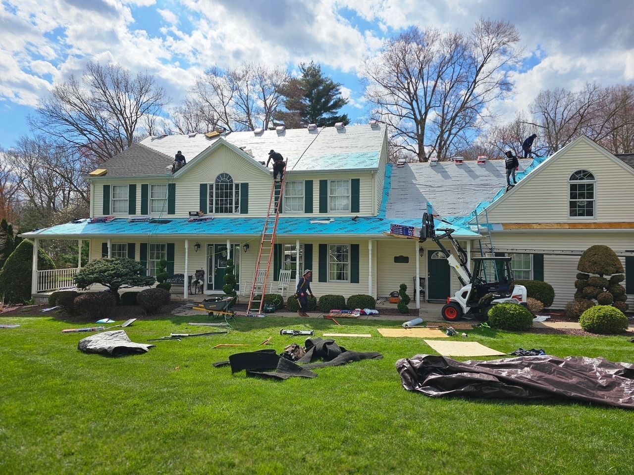Roofers replacing shingles on a two-story white house with green shutters, blue underlayment, and a small Bobcat on the lawn.