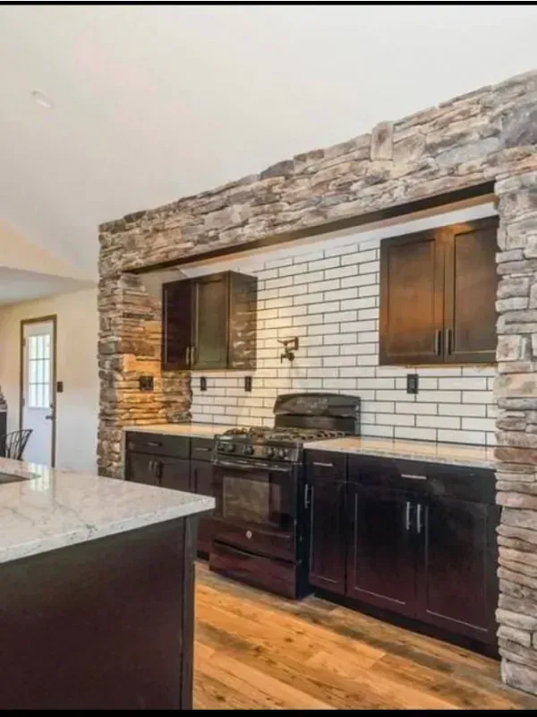 A kitchen with a stove , sink , cabinets and a stone wall.