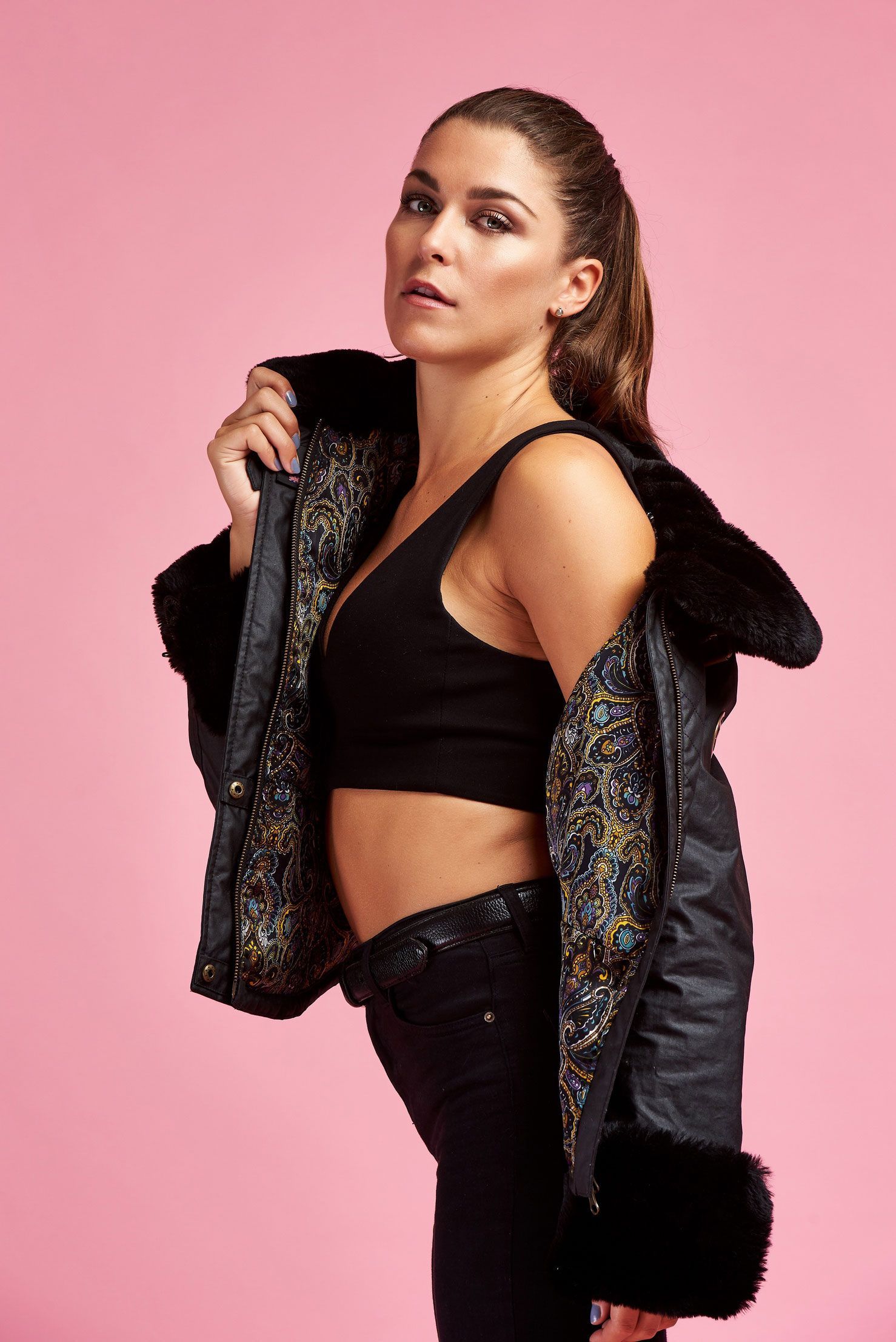 a woman in a black crop top and black jacket is standing on a pink background .