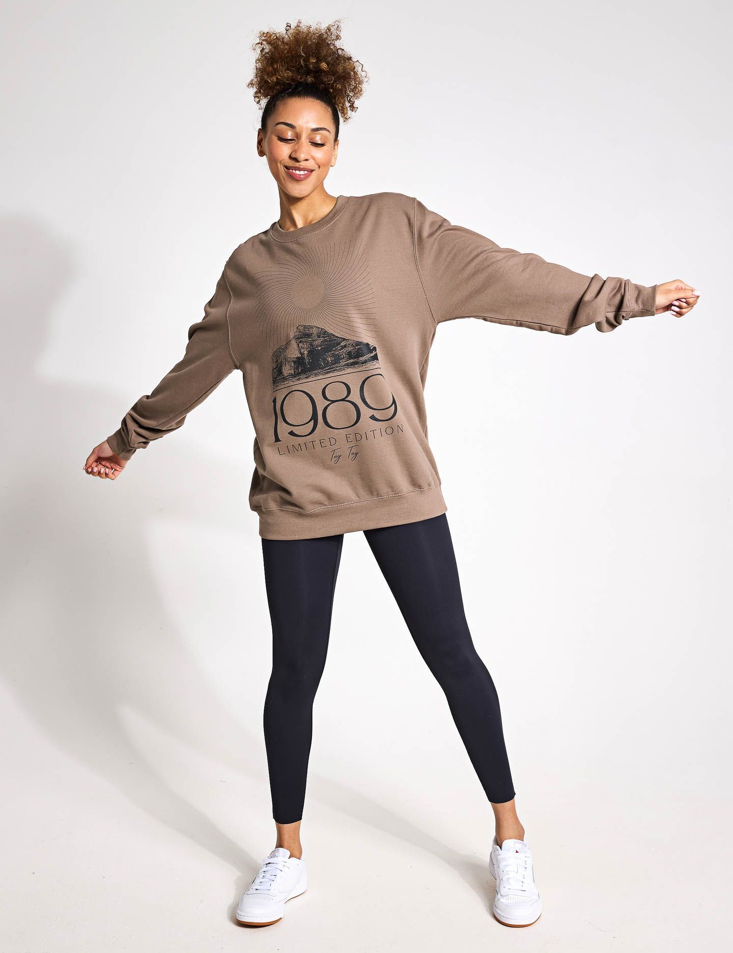 A woman wearing a brown sweatshirt and black leggings is standing with her arms outstretched.