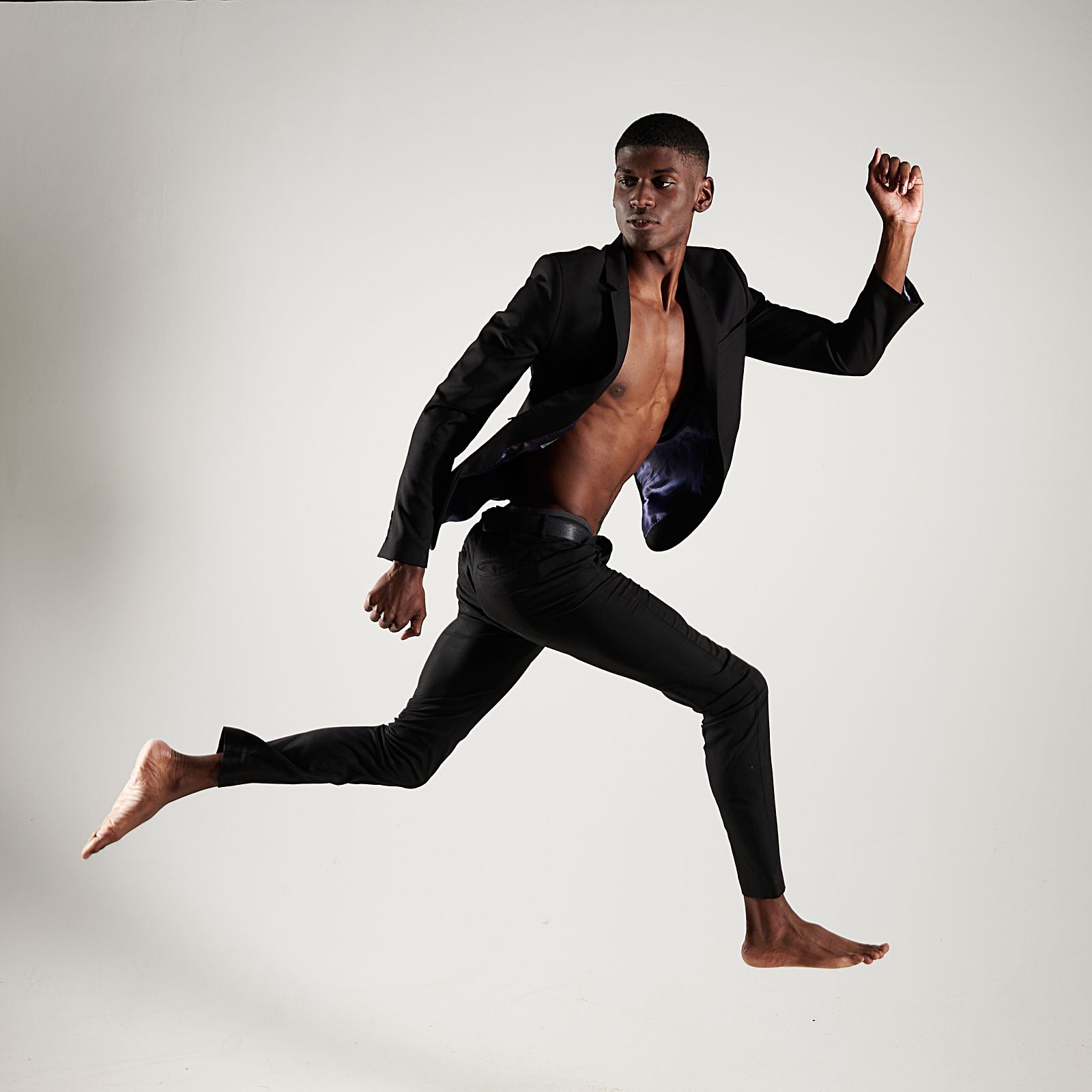 A shirtless man in a black suit is jumping in the air