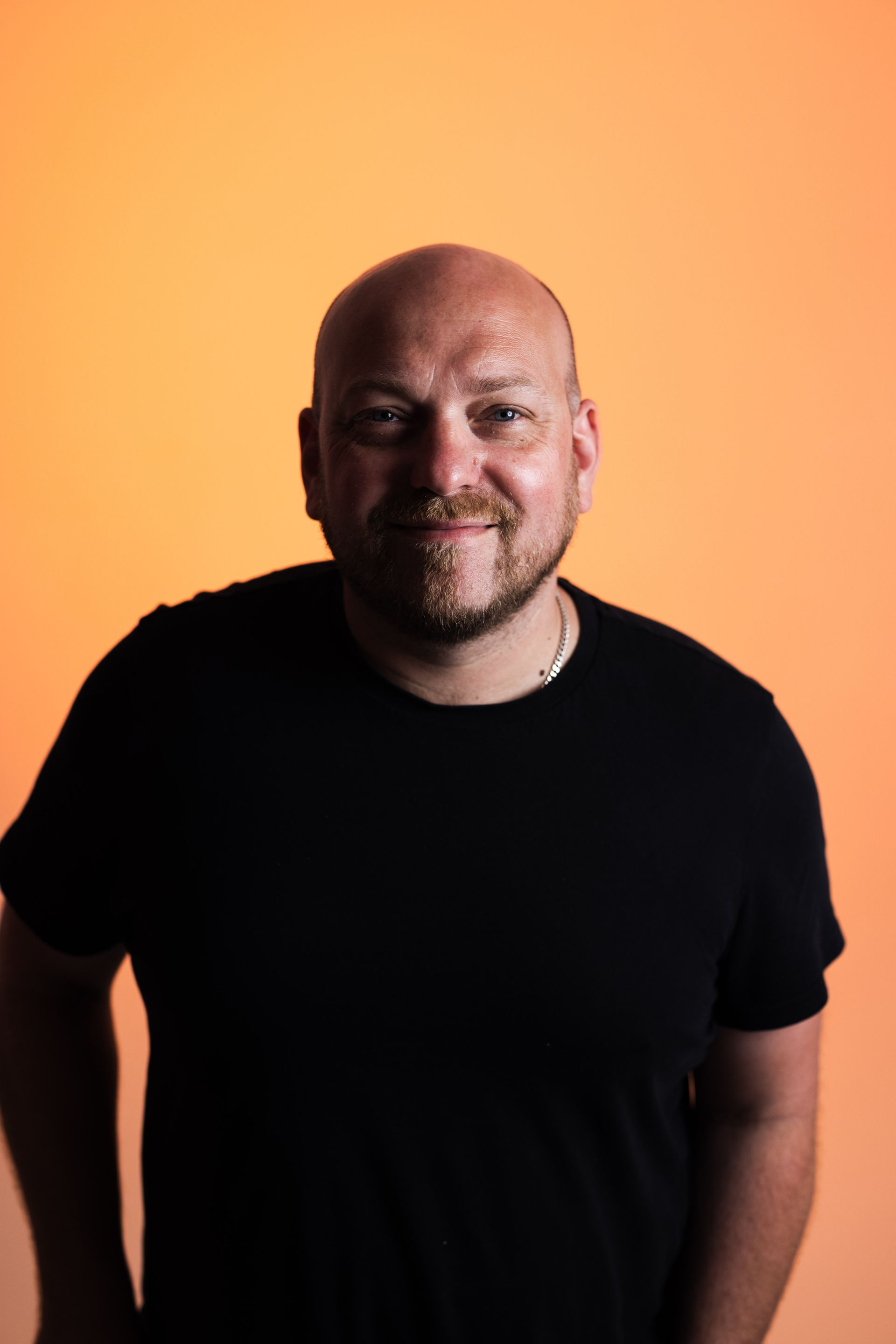 a bald man with a beard is wearing a black shirt and looking at the camera .