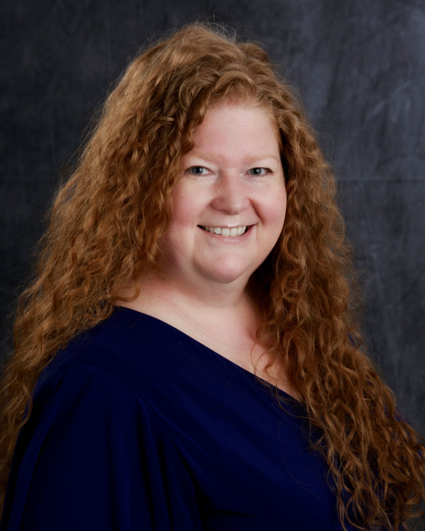Woman with long, curly red hair, smiling in a blue top against a dark background.